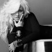 Christina Aguilera con expresión calculadora en el rostro, vistiendo un atuendo negro con brazaletes llamativos en las muñecas y un anillo en su dedo.